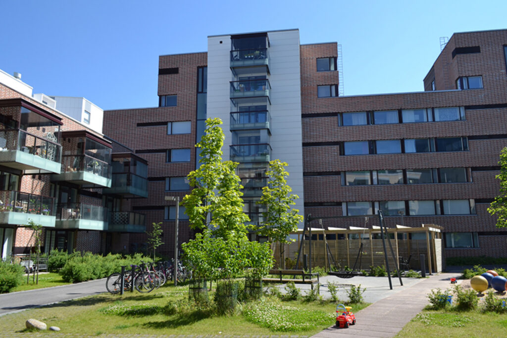 Tummasävyinen kerrostalo, jossa eritasokorkeuksinen katto. Etualalla lasten leikkipaikka, jossa hämähäkkikeinu.