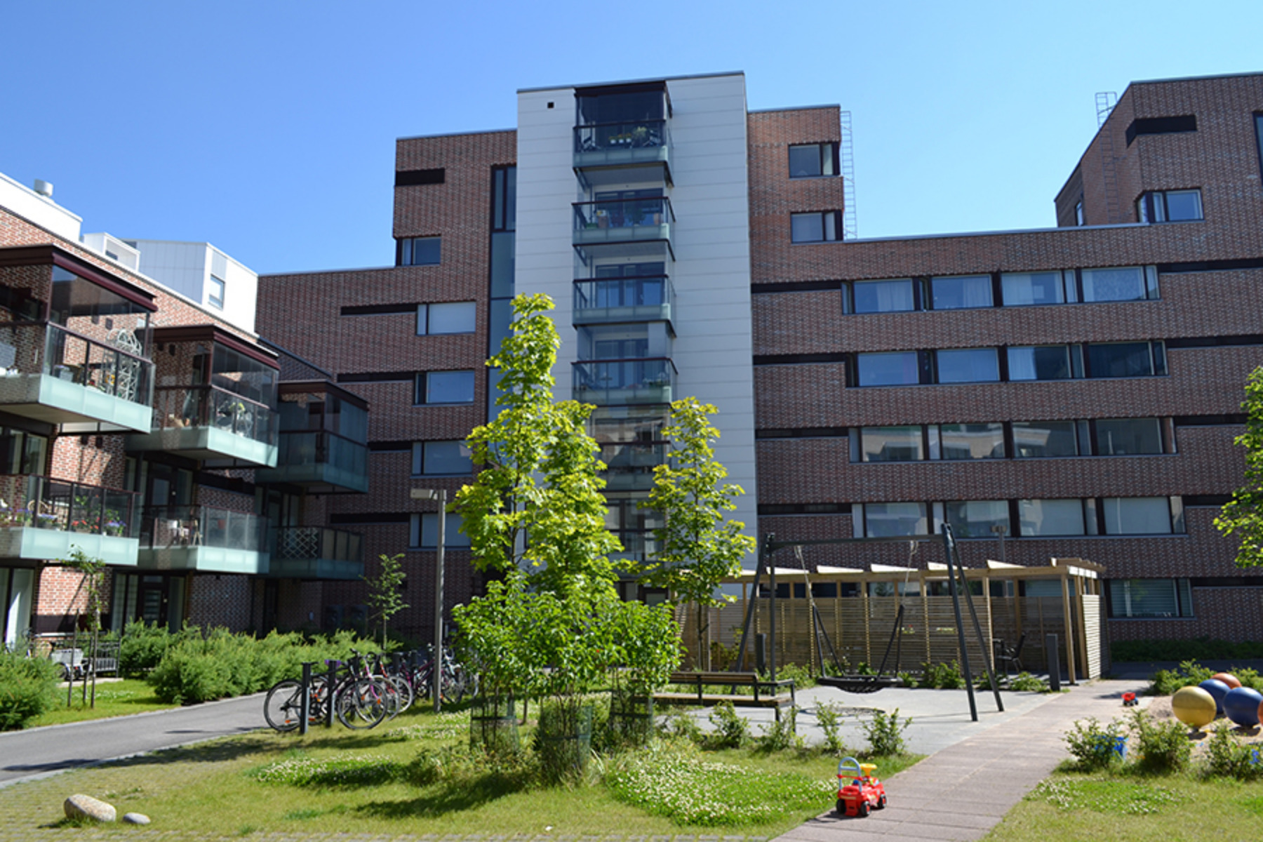 Tummasävyinen kerrostalo, jossa eritasokorkeuksinen katto. Etualalla lasten leikkipaikka, jossa hämähäkkikeinu.